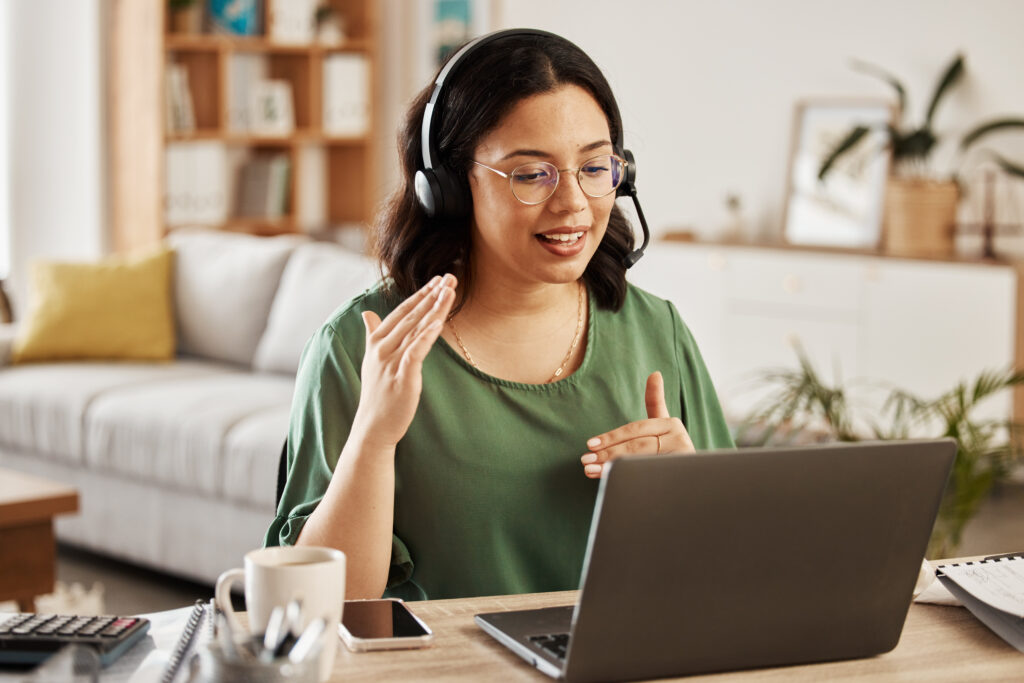 Woman wearing a headset and speaking, seated at a desk with a laptop in a bright home office.