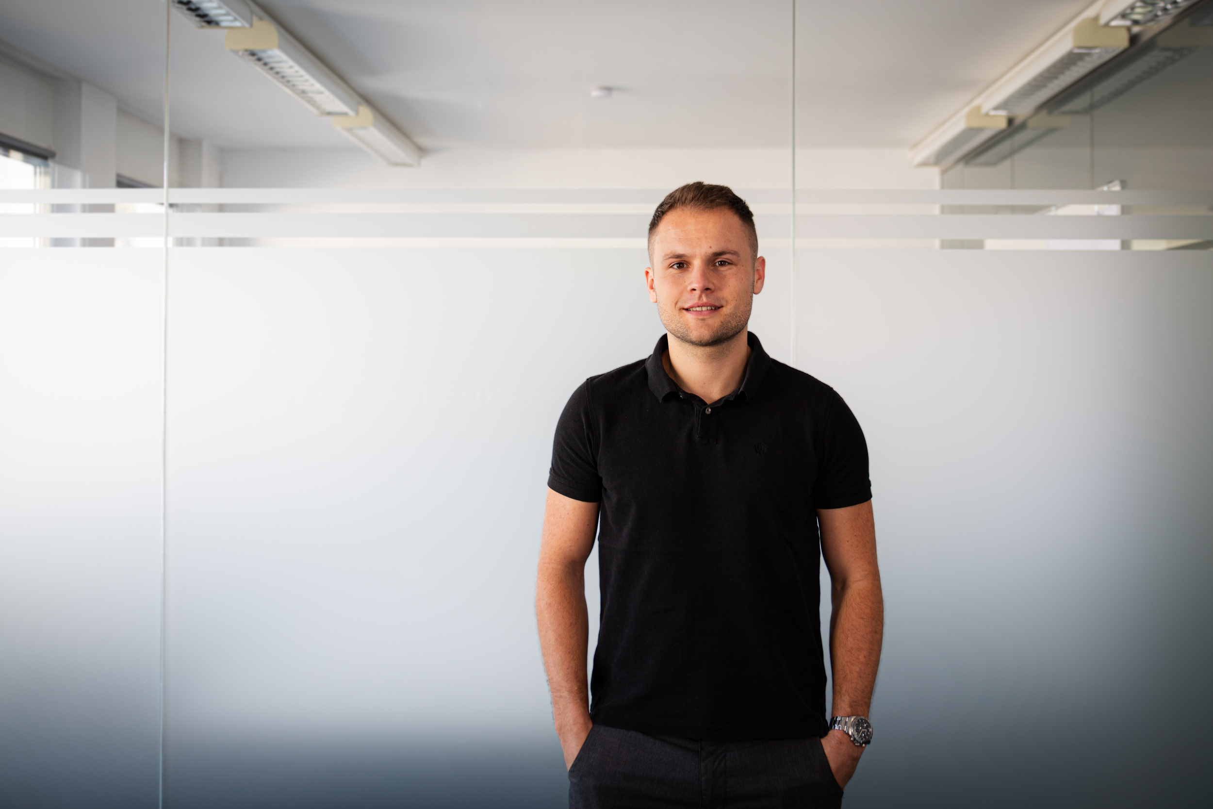 Man in a black polo stands with hands in pockets in a modern office with frosted glass panels behind him.