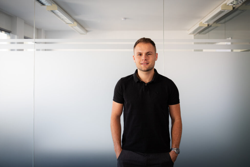 Man in a black polo stands against a glass-paneled office wall, hands in pockets.