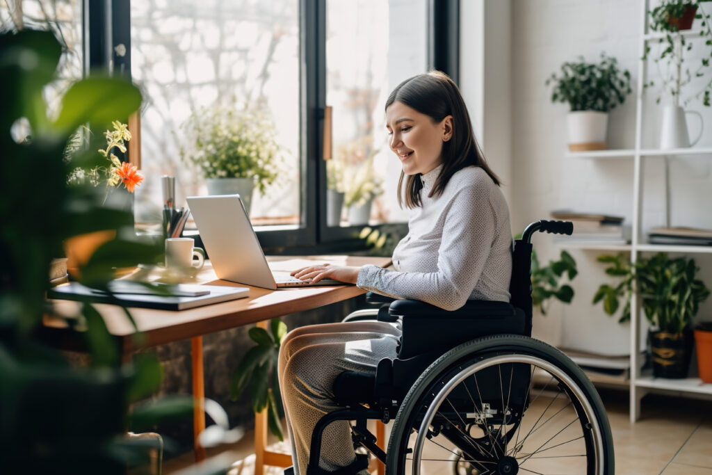 Woman in a wheelchair typing on a laptop at a wooden desk by a sunlit window with plants around her.