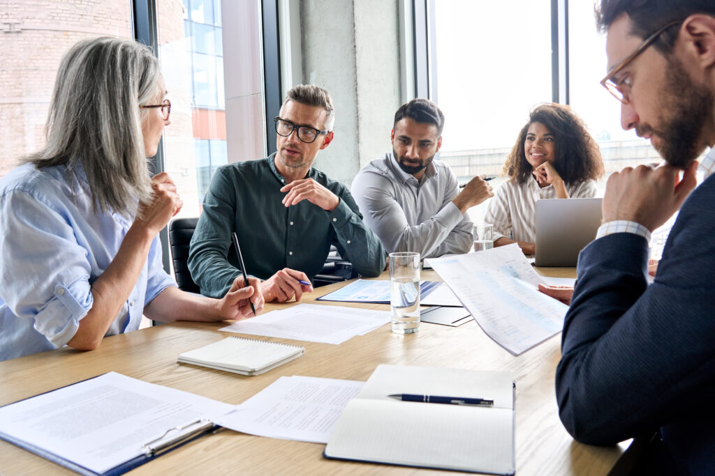 Six professionals sit around a wooden table in a bright office, discussing documents and notes.