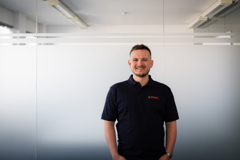 Smiling man in a dark polo shirt standing in an office with frosted glass panels and stripes on the glass.