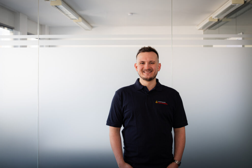 Smiling man in a dark polo shirt stands against a frosted glass office wall with horizontal stripes in the background.