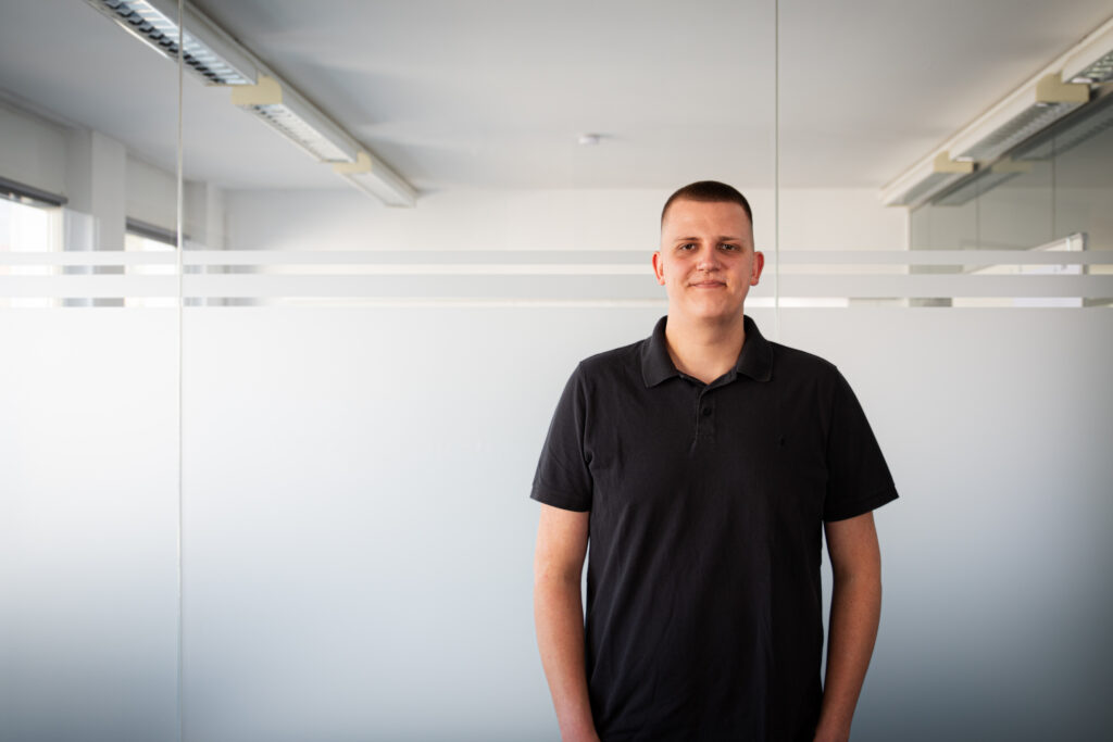 Young man in a black polo shirt standing in a bright office with frosted glass panels behind him.
