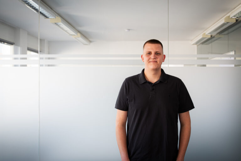 Young man in a black polo shirt standing in a bright office with frosted glass panels behind him.