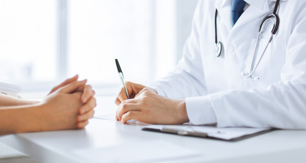 Doctor in white coat with stethoscope signs a document at a desk, assisting a patient.