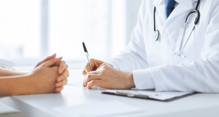 Doctor in white coat with stethoscope signs a document at a desk, assisting a patient.