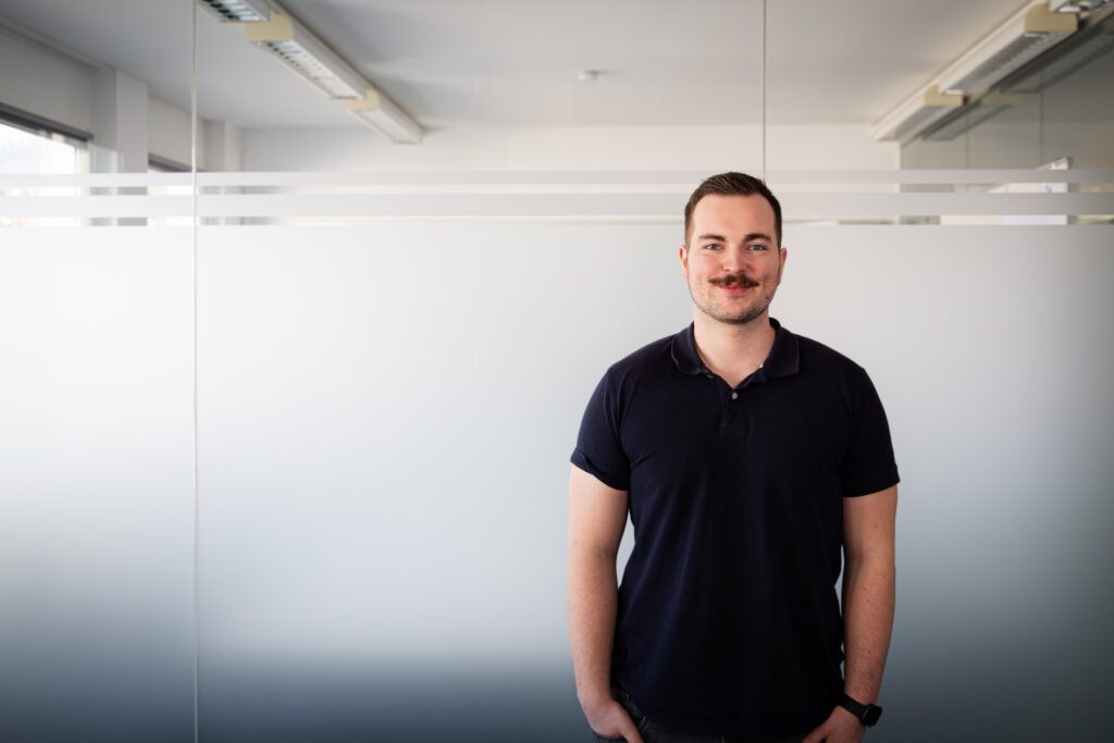 Man wearing a navy polo shirt stands in a bright office with frosted glass panels behind him.