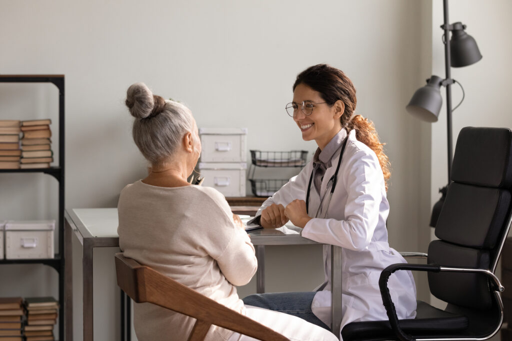 Doctor in a white coat and stethoscope consults an elderly patient across a desk in a clinic, both smiling.
