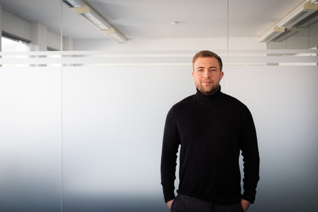 Man in a black turtleneck stands against a glass-walled office with frosted horizontal stripes.