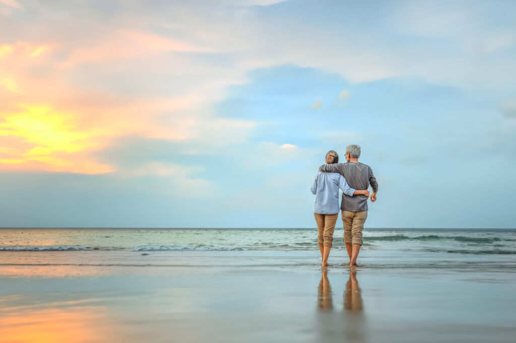 Couple standing on a beach with their arms around each other, watching a sunset over the ocean.