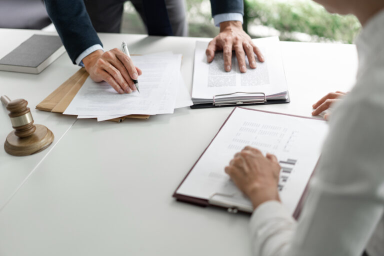 Two people reviewing documents on a white desk; a gavel sits at the edge and a notebook lies nearby.