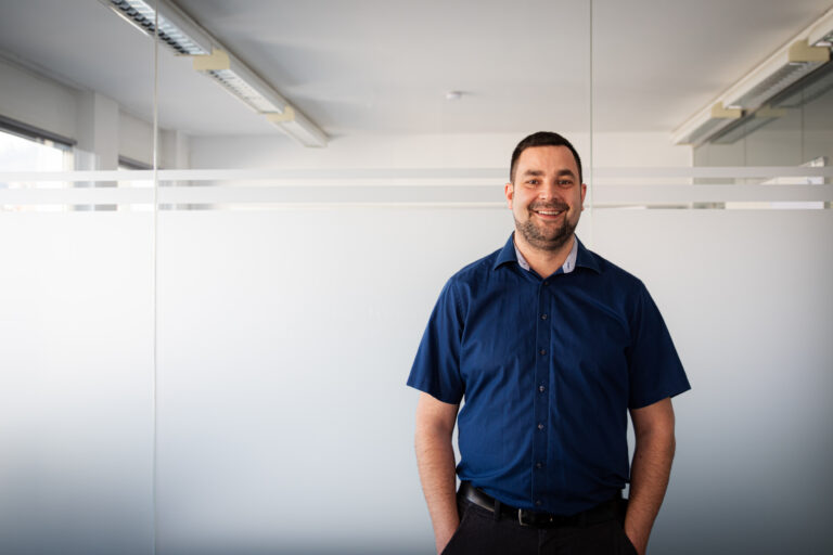 Smiling man in a navy blue shirt stands in a modern office with glass partitions behind him.