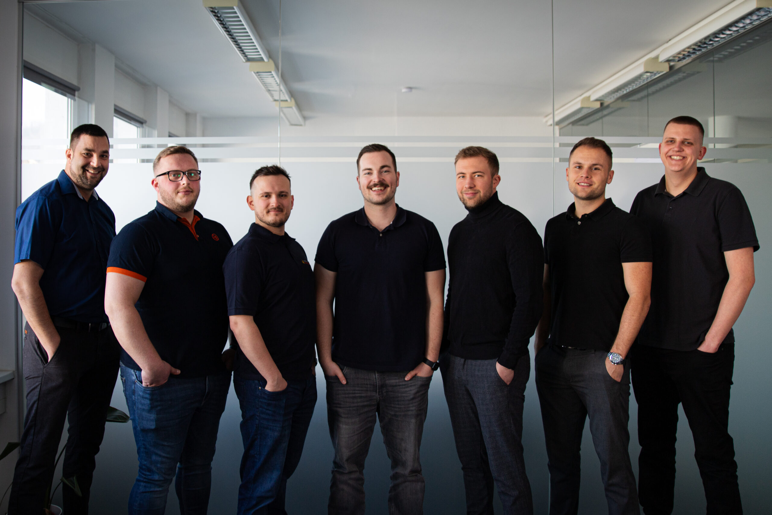 Eight men stand side by side in an office, wearing dark shirts and smiling at the camera in front of a glass partition.