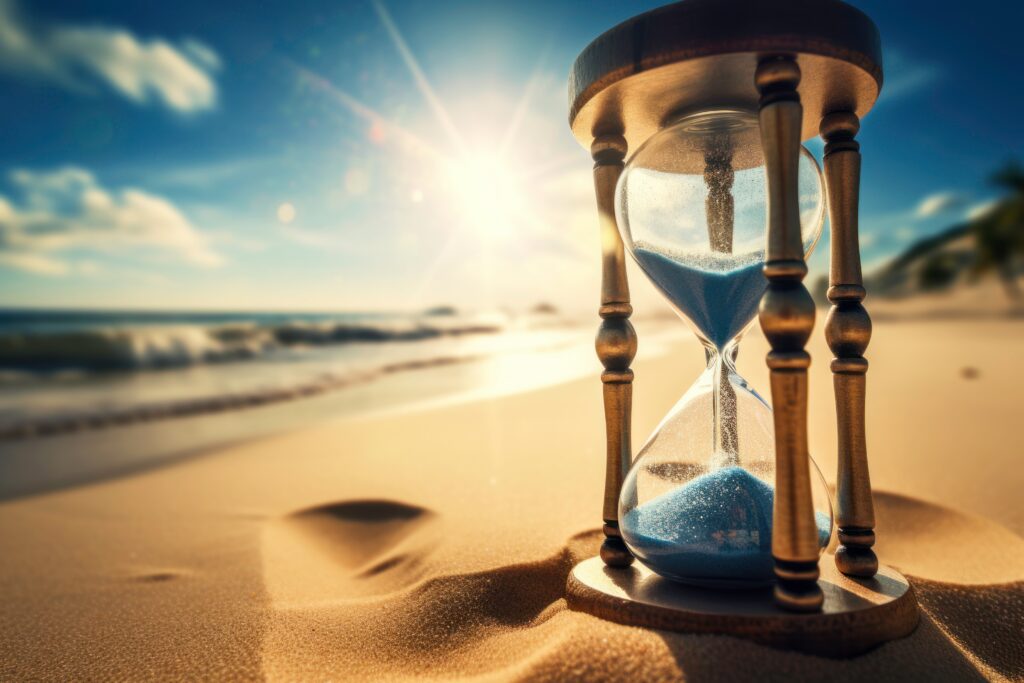 Hourglass on sandy beach with bright sun flare and blue sky in the background.