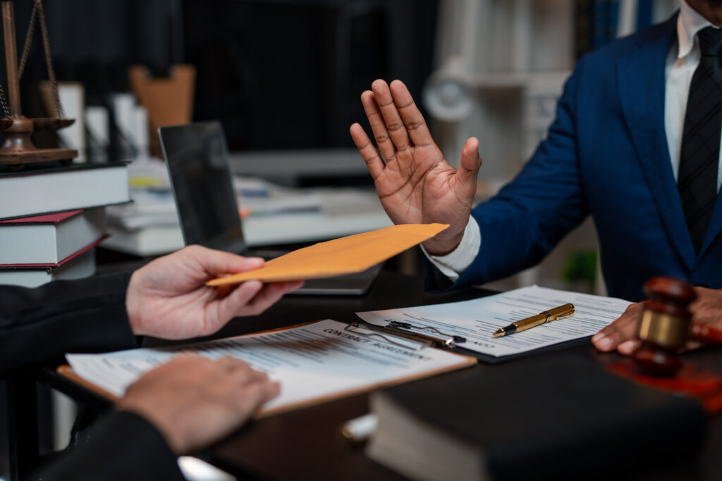Law office scene: A person in a blue suit hands an orange envelope across a desk with legal papers and a laptop nearby, scales in the background.