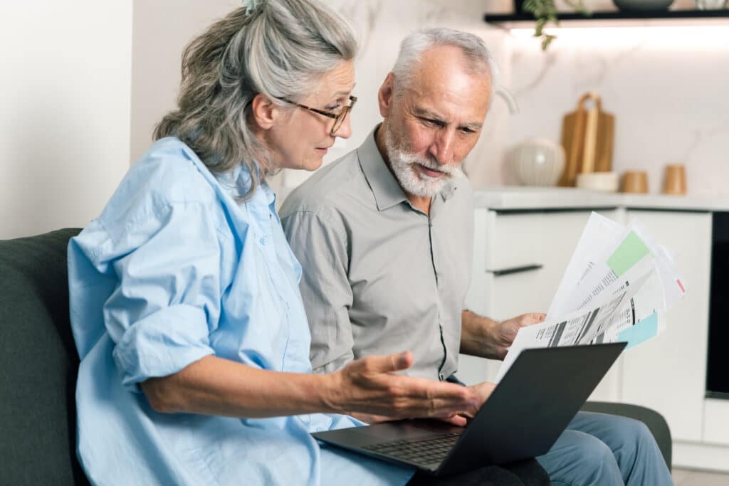 A husband and wife sit on a couch looking at utility bills and electricity, gas or heating statements on their laptop, analyzing family budget and discussing finances together at home.