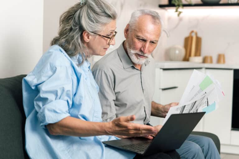 A husband and wife sit on a couch looking at utility bills and electricity, gas or heating statements on their laptop, analyzing family budget and discussing finances together at home.